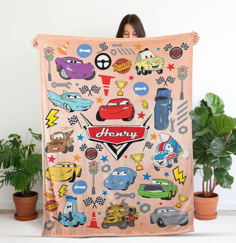 May include: A light peach-colored fleece blanket featuring a repeating pattern of various cartoon cars, traffic lights, checkered flags, and tools from the movie Cars. The center displays a large red emblem with the name Henry written in white.