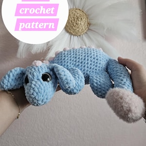 May include: A light blue crocheted dragon with pink accents and large black eyes. The dragon is lying on its belly with its head turned to the side. The text "PDF crochet pattern" is visible in the upper left corner of the image.