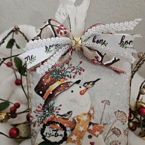 May include: A wooden Christmas ornament featuring a snowman design. The snowman wears a hat, scarf, and has birds perched on it. The ornament has a ribbon with the word "home" and a small gold bell. The ornament is decorated with a bow.