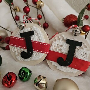 May include: Two round Christmas ornaments with the letter "J" in black, accented with red ribbon and white lace. The ornaments are surrounded by red berries, bells, and other festive decorations, creating a holiday theme.