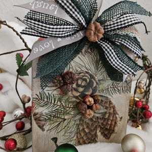 May include: Wooden house-shaped decoration with a pine cone and evergreen design. The decoration is adorned with a large bow made of green and black checkered ribbon, and gray ribbon with the words "Love" and "Laugh". Includes green and red Christmas ornaments.