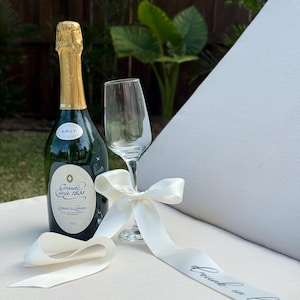 May include: A bottle of Brut champagne with a gold foil top, a champagne flute, and a white ribbon with the text "drunk in love". The bottle has a green label with gold lettering. The ribbon is tied in a bow.