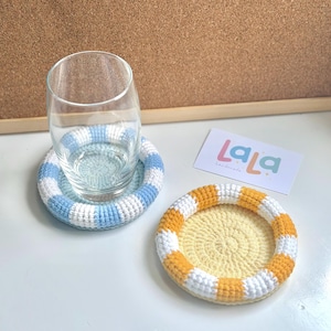 Peut inclure: Deux dessous de verre crochetés à la main. L'un a une bordure rayée bleue et blanche avec un verre dessus. L'autre a une bordure rayée orange et blanche avec un centre jaune. Une carte de visite est également présente.