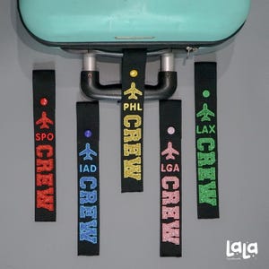 May include: Black luggage tags with the word "CREW" in various colors, including red, blue, gold, pink, and green. Each tag features an airplane graphic and an airport code like LAX, PHL, and IAD. The tags are hanging from a teal suitcase.