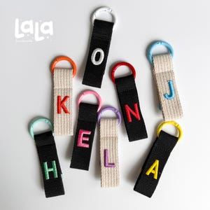 May include: A collection of fabric keychains featuring black or beige webbing straps with embroidered letters in various colors. Each strap is attached to a colorful plastic ring. The text "LaLa handmade" appears in the top left corner.
