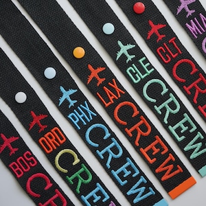 May include: Black luggage straps embroidered with airport codes and the word "CREW" in various colors. Each strap features a small airplane icon and a colorful pin.