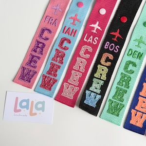 May include: A collection of colorful fabric luggage tags with the word "CREW" in glittery letters. Each tag features a different airport code and a small embroidered airplane. The tags are in various colors, including pink, blue, and black.