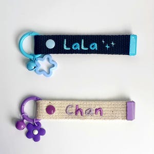 May include: Two personalised keychains. The top keychain is black with the name "LaLa" embroidered in teal, a blue charm, and a star-shaped charm. The bottom keychain is beige with the name "Chan" embroidered in purple, a purple flower charm, and a purple charm.