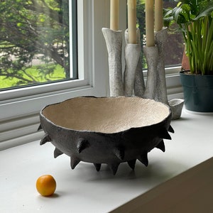 May include: A black bowl with a textured, spiky exterior and a cream-colored interior. The bowl is sitting on a white surface next to a small orange fruit.