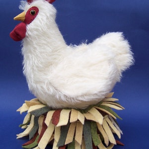 May include: A white fluffy chicken with a red comb and wattle sits on a nest of brown, green, and beige fabric strips.