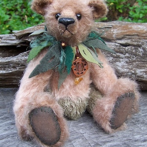 May include: A light brown teddy bear with a green and brown leafy collar and a wooden pendant. The bear is sitting on a wooden surface.