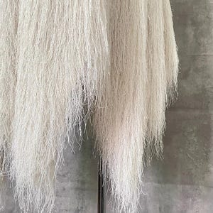 May include: Close-up of a white, textured, hanging art piece. The artwork is composed of numerous thin, thread-like strands cascading downwards. The strands are a soft white colour, creating a delicate and airy aesthetic against a grey background.