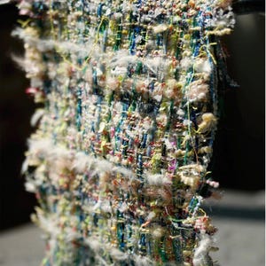 Op de afbeelding: Close-up van een kleurrijk, getextureerd stofstaal. Het textiel heeft een mix van blauwe, groene, roze en witte draden die door elkaar zijn geweven. De randen hebben een gerafelde, onafgewerkte uitstraling, met losse draden en pluizige witte accenten.