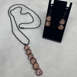 May include: A jewelry set consisting of a necklace and a pair of drop earrings. The necklace features a black ball chain with a vertical pendant of six round, light pink crackle glass beads wrapped in black wire. The earrings match the pendant design.