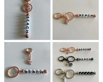 May include: Rose gold keychains with the word "BEACH" spelled out in white and black letter beads. Each keychain has a lobster clasp and a charm, including a starfish and a dolphin.