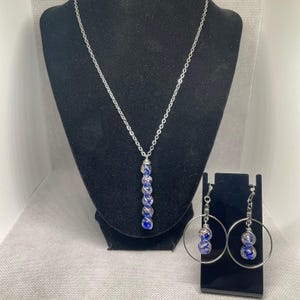 May include: A silver-toned necklace featuring a vertical pendant of five spherical blue and clear glass beads wrapped in silver wire, accompanied by matching hoop earrings, each containing two identical blue and clear glass beads.