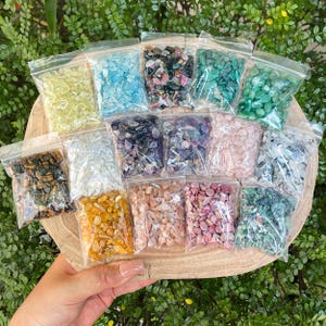 Puede incluir: Una colección de pequeñas piedras preciosas de colores en bolsas de plástico transparente, dispuestas sobre una bandeja de madera. Las piedras preciosas varían en color, incluyendo amarillo, azul, verde, morado, rosa y marrón. La bandeja se sostiene contra un fondo de follaje verde.