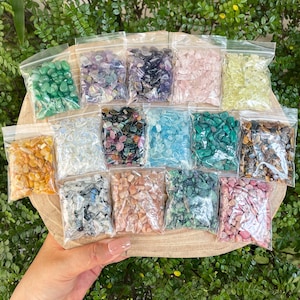 May include: A selection of small, colourful gemstones in clear plastic bags. The gemstones are various colours, including green, purple, pink, and yellow. The bags are arranged on a wooden surface, with a hand holding the wooden surface.