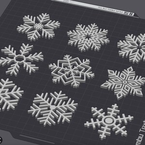 Puede incluir: Una colección de nueve diseños de copos de nieve blancos, cada uno con patrones y formas únicos. Los copos de nieve están dispuestos sobre una superficie texturizada de color gris oscuro, mostrando sus intrincados detalles. La imagen está bien iluminada.