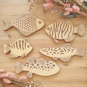 May include: Five wooden fish cutouts with etched details, arranged on a light wood surface. Each fish has a unique design, including patterns of lines, dots, and scales. The fish vary in shape and size, with a natural wood color.