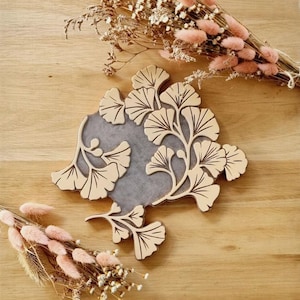 May include: A wooden coaster with a ginkgo leaf design. The coaster is light brown with a gray center. The ginkgo leaves are intricately carved with dark brown lines. Dried flowers in shades of pink and beige are arranged around the coaster on a wooden surface.