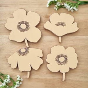 Puede incluir: Cuatro posavasos de cuero beige con forma de flor, con centros y tallos marrones. Los posavasos están dispuestos sobre una superficie de madera clara, con pequeñas flores blancas en el fondo. Un accesorio decorativo para el hogar.