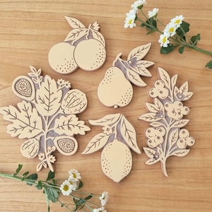 Puede incluir: Una colección de recortes de madera con forma de fruta, que incluyen naranjas, una pera, un limón, higos y bayas. Los recortes son de color beige claro con detalles grabados en marrón oscuro y están dispuestos sobre una superficie de madera. Flores blancas en el fondo.