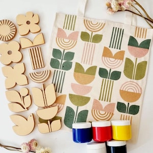 Puede incluir: Un conjunto de formas de madera, incluyendo flores y formas geométricas, junto a una bolsa de lona con un estampado floral. La imagen también presenta pequeños botes de pintura azul, roja, amarilla, blanca y negra, sugiriendo un proyecto de manualidades.