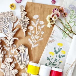 May include: A collection of wooden floral stamps, fabric tote bags, and paint jars. The stamps feature various botanical designs. The tote bags display printed floral patterns in white, yellow, and green. Jars of yellow, white, and red paint are also visible.