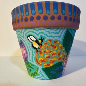 Può includere: Un vaso di fiori in terracotta dipinto a mano con un motivo floreale blu, verde, giallo, arancione e rosa. Un'ape è dipinta sul lato del vaso.
