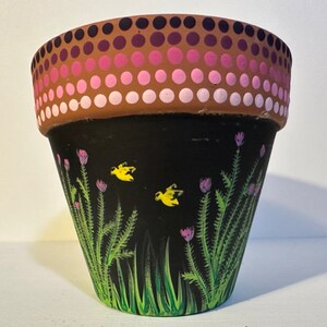 Puede incluir: Una maceta de terracota con fondo negro y un diseño de lunares rosa y blanco alrededor del borde superior. La maceta está pintada con hierba verde y flores rosas con mariposas amarillas.