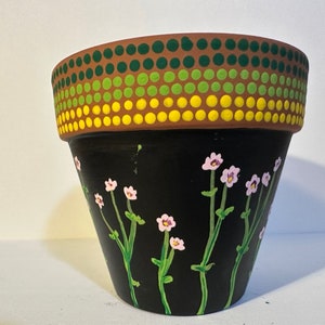 Puede incluir: Una maceta de terracota negra con un diseño pintado de flores blancas y tallos verdes. El borde superior de la maceta está decorado con un patrón de puntos multicolores en tonos marrones, verdes y amarillos.