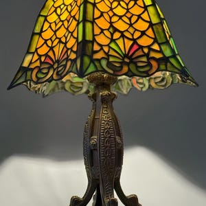 Op de afbeelding: Een tafellamp met een vierkante glas-in-loodkap met een geometrisch patroon in geel-, oranje- en groentinten. De lamp heeft een donkerbruine, getextureerde metalen voet met gedetailleerde reli&euml;fversieringen en een vierpotig ontwerp.