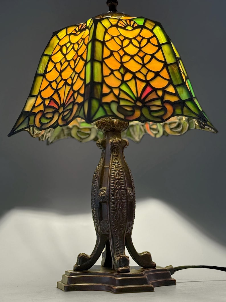 Op de afbeelding: Een tafellamp met een vierkante glas-in-loodkap met een geometrisch patroon in geel-, oranje- en groentinten. De lamp heeft een donkerbruine, getextureerde metalen voet met gedetailleerde reli&euml;fversieringen en een vierpotig ontwerp.