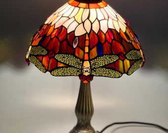 Op de afbeelding: Een glas-in-lood tafellamp met een koepelvormige kap voorzien van een libellenmotief in oranje, rode en gele tinten. De lamp heeft een donkerbronskleurige metalen voet met een gegroefde steel en een decoratieve, reliëfrijke ronde voet.