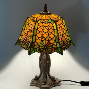 May include: A stained glass table lamp with a bronze base. The lampshade is a vibrant mix of yellow, orange, green, and red glass pieces, creating a mosaic pattern. The base has an ornate, detailed design.