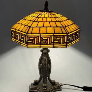 Op de afbeelding: Een tafellamp met een zeshoekige kap van gebrandschilderd glas in geel en amber, voorzien van een geometrisch Grieks sleutelpatroon. De lamp heeft een donkerbronskleurige metalen voet met gedetailleerde texturen en een driepotig ontwerp.
