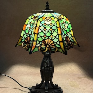Op de afbeelding: Een tafellamp met een glas-in-loodkap met een geschulpte rand, voorzien van een patroon van groene, oranje en gele glassegmenten. De lamp heeft een donkere, sierlijke metalen voet met een gestructureerde steel en een vierkante voetplaat.