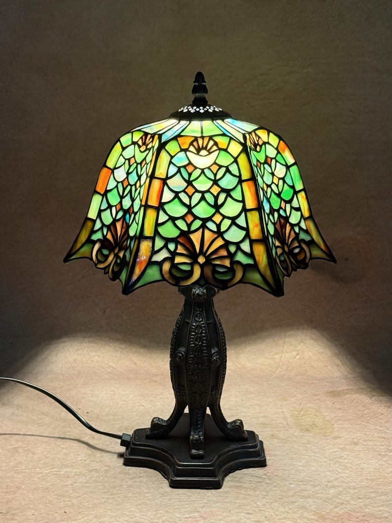 Op de afbeelding: Een tafellamp met een glas-in-loodkap met een geschulpte rand, voorzien van een patroon van groene, oranje en gele glassegmenten. De lamp heeft een donkere, sierlijke metalen voet met een gestructureerde steel en een vierkante voetplaat.