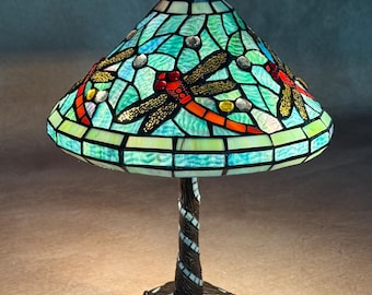 Op de afbeelding: Een Tiffany-stijl tafellamp met een Tiffany-glas lampenkap met een libellenmotief. De lampenkap is een mix van blauw, groen, rood en goud glas. De voet is van gedraaid, donkerbruin metaal met een mozaïekpatroon.