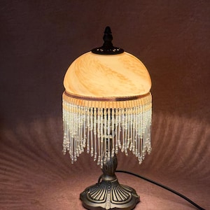 May include: A decorative table lamp with a cream-colored, marbled glass shade and a beaded fringe. The lamp has a bronze-colored base and a black power cord. The lamp is designed in a vintage style.