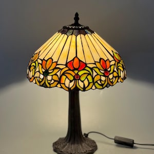 May include: A stained glass table lamp with a bronze base. The lampshade features a floral design with yellow, orange, and green glass panels. The lamp has a pull chain and a cord with a switch. The lamp is approximately 18 inches tall.