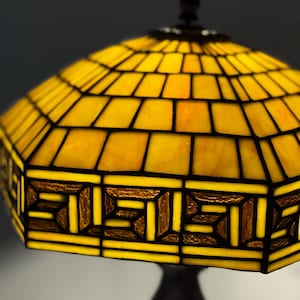 Op de afbeelding: Een glas-in-lood lampenkap met een levendig geel geometrisch patroon en donkere loden lijnen. De onderste rand toont een herhalend rechthoekig Grieks sleutelmotief in getextureerd amberkleurig glas, contrasterend met de heldergele panelen.