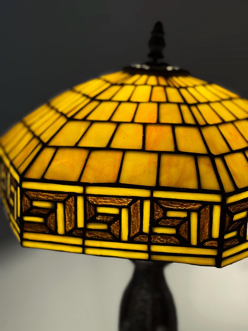 Op de afbeelding: Een glas-in-lood lampenkap met een levendig geel geometrisch patroon en donkere loden lijnen. De onderste rand toont een herhalend rechthoekig Grieks sleutelmotief in getextureerd amberkleurig glas, contrasterend met de heldergele panelen.