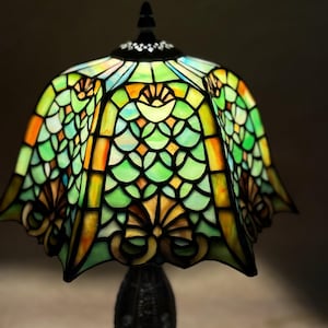 Op de afbeelding: Een glas-in-loodlamp met een geschulpte kap met een herhalend schubpatroon in tinten groen, geel en oranje. De glaspanelen zijn verbonden door donkere metalen lijnen en de kap is afgewerkt met een zwarte knop aan de bovenzijde.