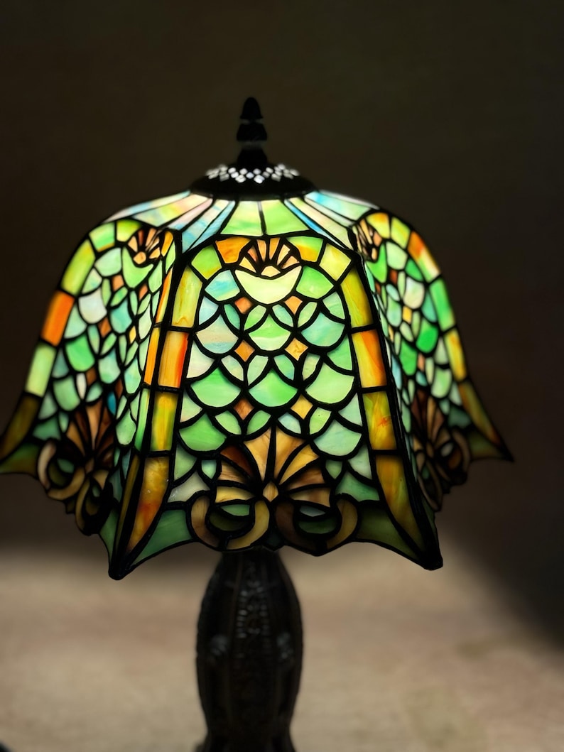 Op de afbeelding: Een glas-in-loodlamp met een geschulpte kap met een herhalend schubpatroon in tinten groen, geel en oranje. De glaspanelen zijn verbonden door donkere metalen lijnen en de kap is afgewerkt met een zwarte knop aan de bovenzijde.