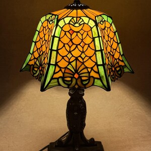 Op de afbeelding: Een tafellamp met een glas-in-lood kap met een geschulpte rand, voorzien van een levendig patroon van oranje schubvormige figuren en groene accenten. De lamp rust op een donkere, getextureerde metalen voet met een sierlijk ontwerp.