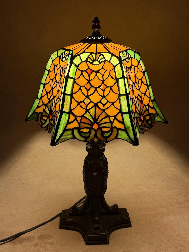 Op de afbeelding: Een tafellamp met een glas-in-lood kap met een geschulpte rand, voorzien van een levendig patroon van oranje schubvormige figuren en groene accenten. De lamp rust op een donkere, getextureerde metalen voet met een sierlijk ontwerp.