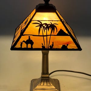 May include: A decorative table lamp with a stained glass shade depicting a desert scene with palm trees, a camel, and a pyramid. The lamp has a bronze-coloured base and a black finial. The shade glows with warm orange and yellow hues when lit.