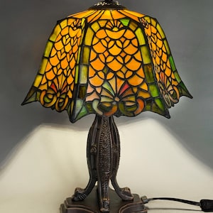 Op de afbeelding: Een glas-in-lood tafellamp met een uitlopende kap voorzien van een geometrisch schubpatroon in levendige oranje en groene tinten. De lamp heeft een donkere, getextureerde metalen voet met een decoratieve steel en een vierkante voet.